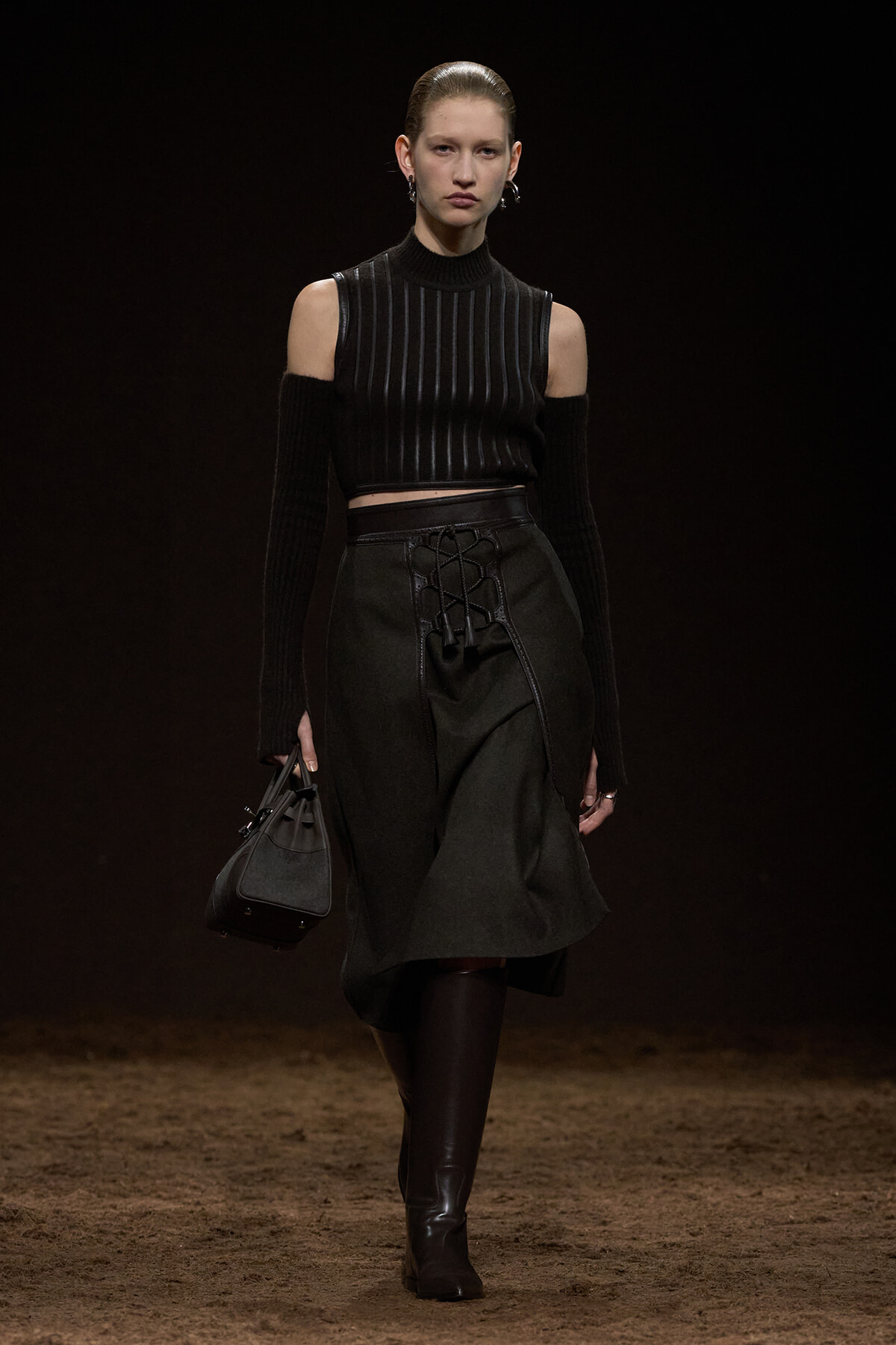 Runway model in an all-black outfit: cold-shoulder knit top, lace-up waist skirt, knee-high boots, and a small handbag.