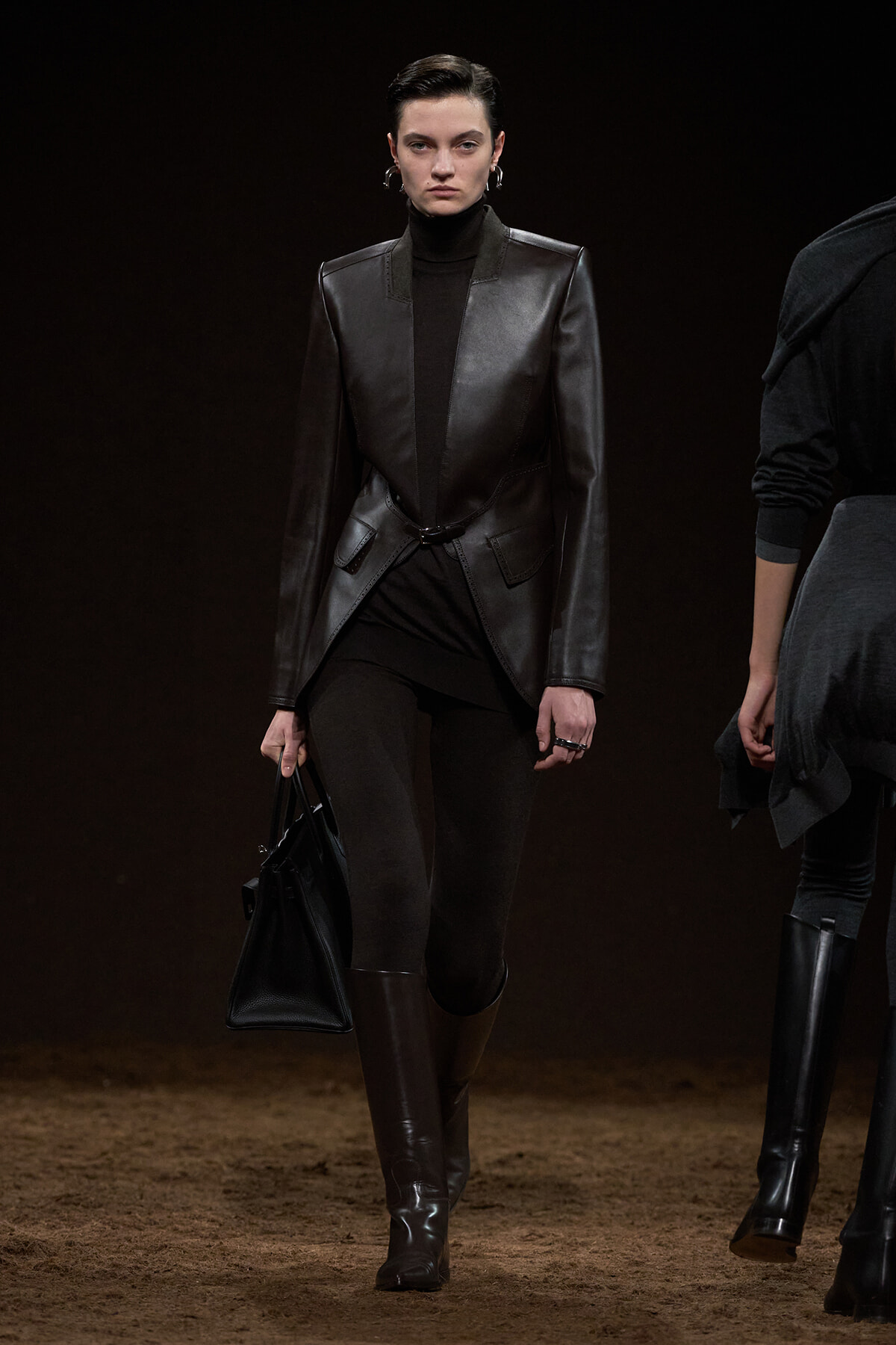 Runway model in a black leather blazer over a turtleneck, black pants, and tall brown boots carrying a black handbag.