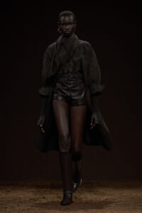 Model walking a dim runway in a dark, layered outfit: oversized cardigan, leather shorts, and knee-high boots.