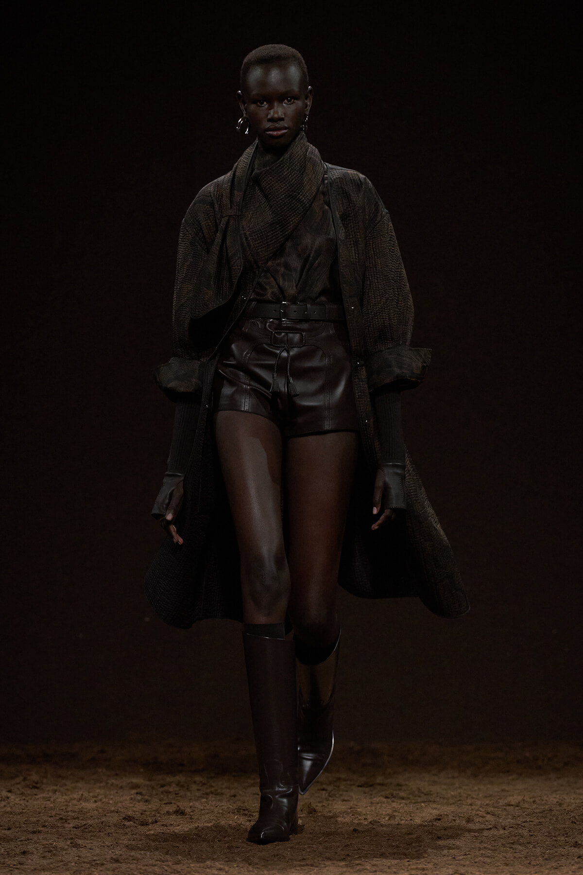 Model walking a dim runway in a dark, layered outfit: oversized cardigan, leather shorts, and knee-high boots.