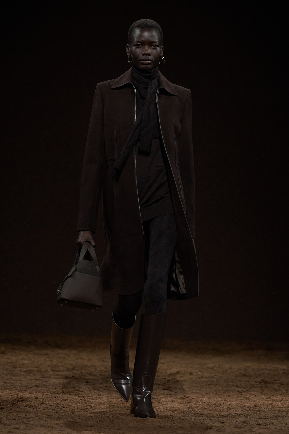 Model walking a dimly lit runway in a long dark brown suede coat, black scarf, and knee-high boots, carrying a gray handbag.