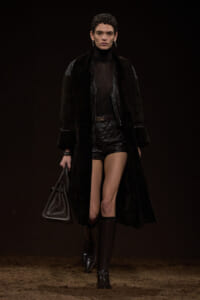 Female model walking a dark runway in an all-black outfit: long fur-cur coat over a sheer black top, quilted leather shorts, belt, tall boots, and a black handbag.