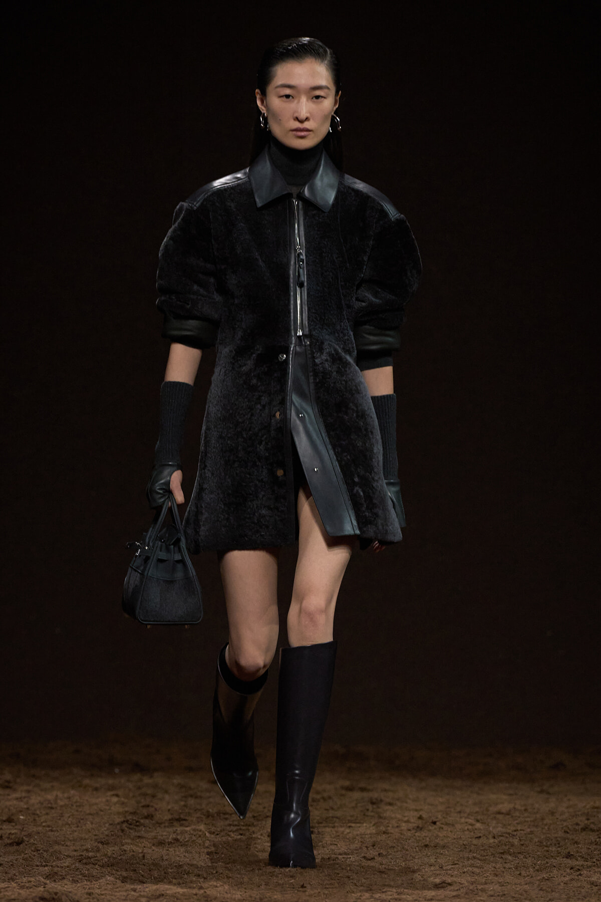Model on a runway wearing a black textured coat with leather collar, matching gloves, knee-high boots, and a small handbag.