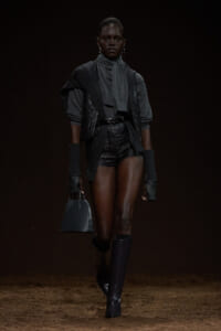 Runway model in a dark, layered charcoal outfit with a quilted vest, scarf, long gloves, and knee-high boots, carrying a small black handbag.