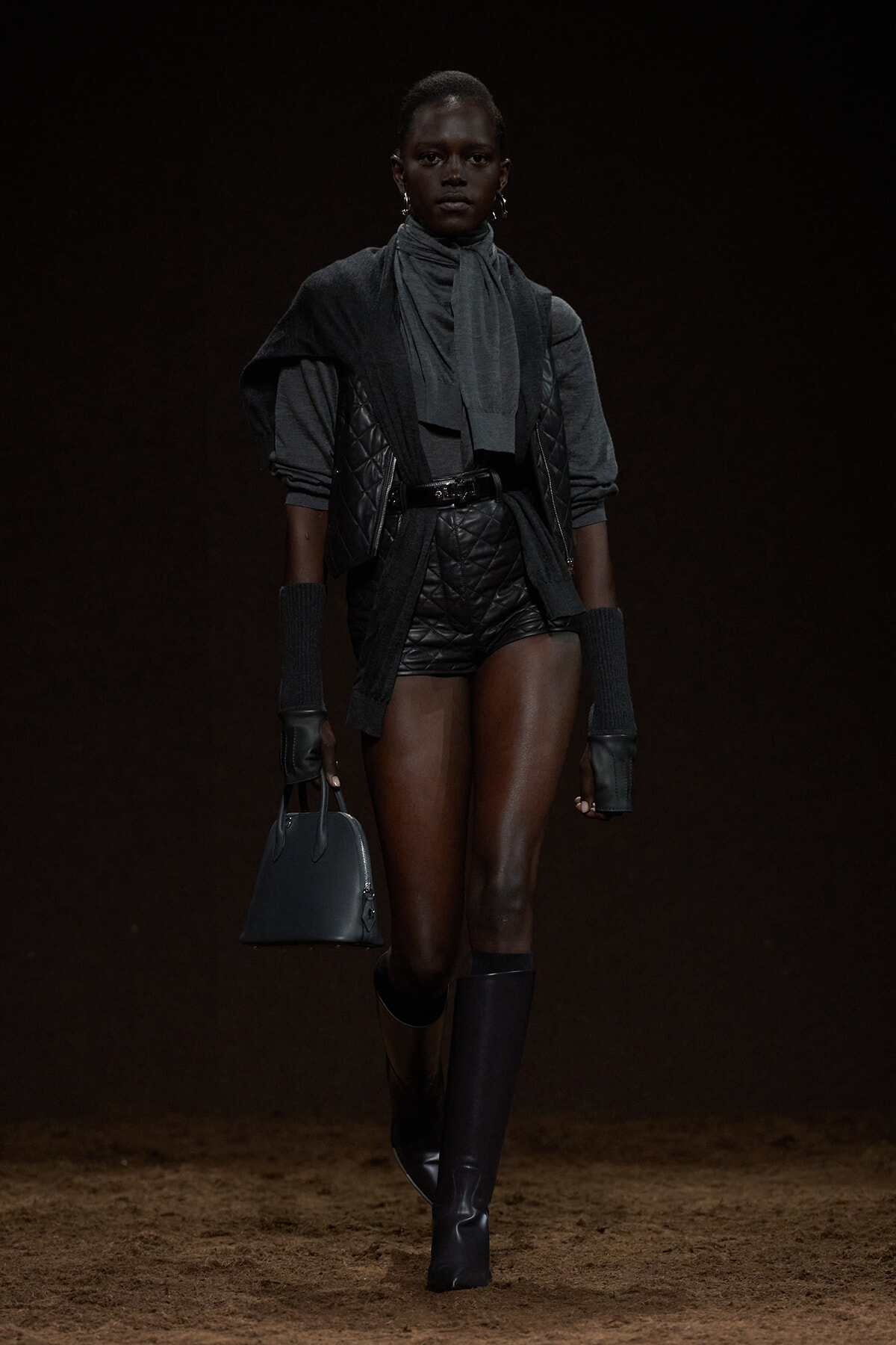Runway model in a dark, layered charcoal outfit with a quilted vest, scarf, long gloves, and knee-high boots, carrying a small black handbag.