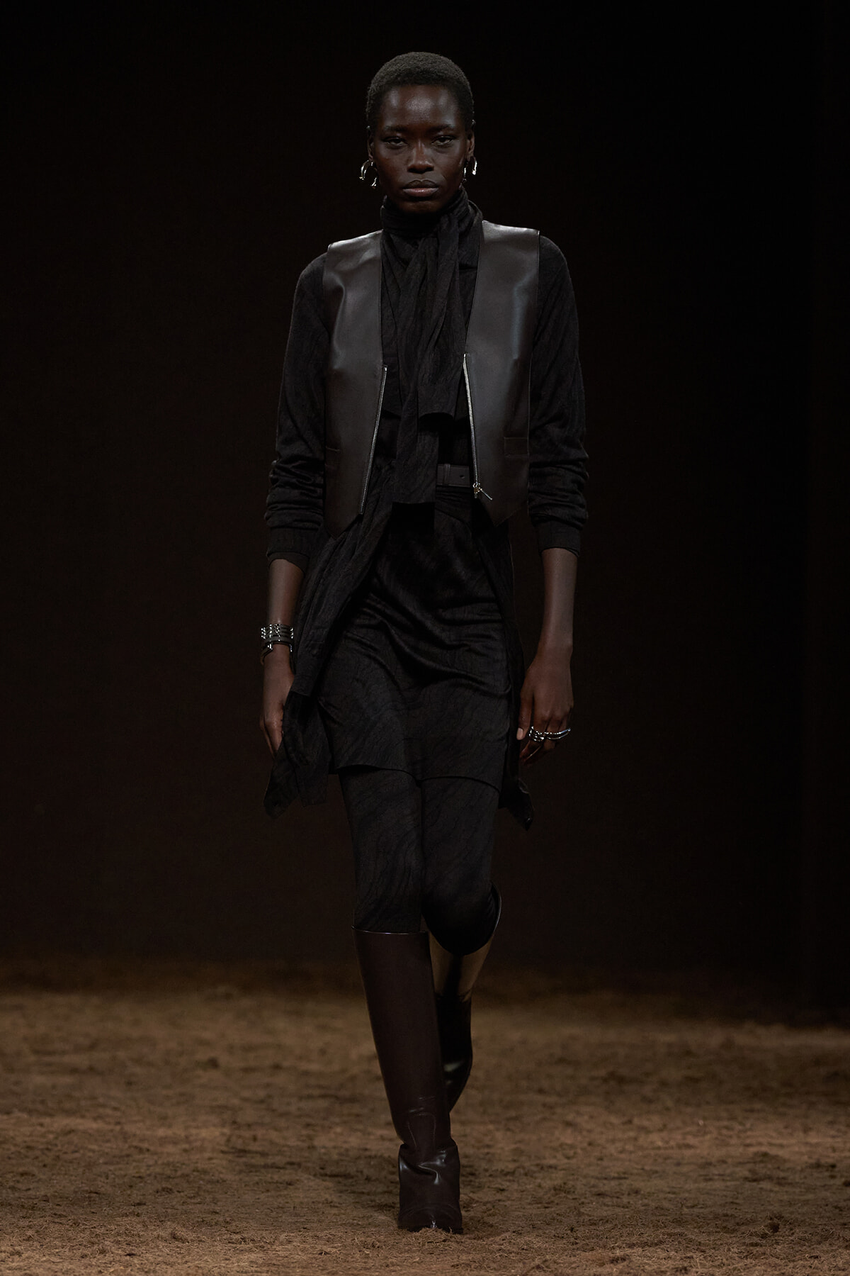 Model walking a runway in a black layered ensemble: long dress, leather vest, and tall boots against a dark background.