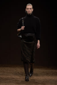 Model walking on a dim runway in a black turtleneck, textured midi skirt, knee-high boots, and a black shoulder bag.