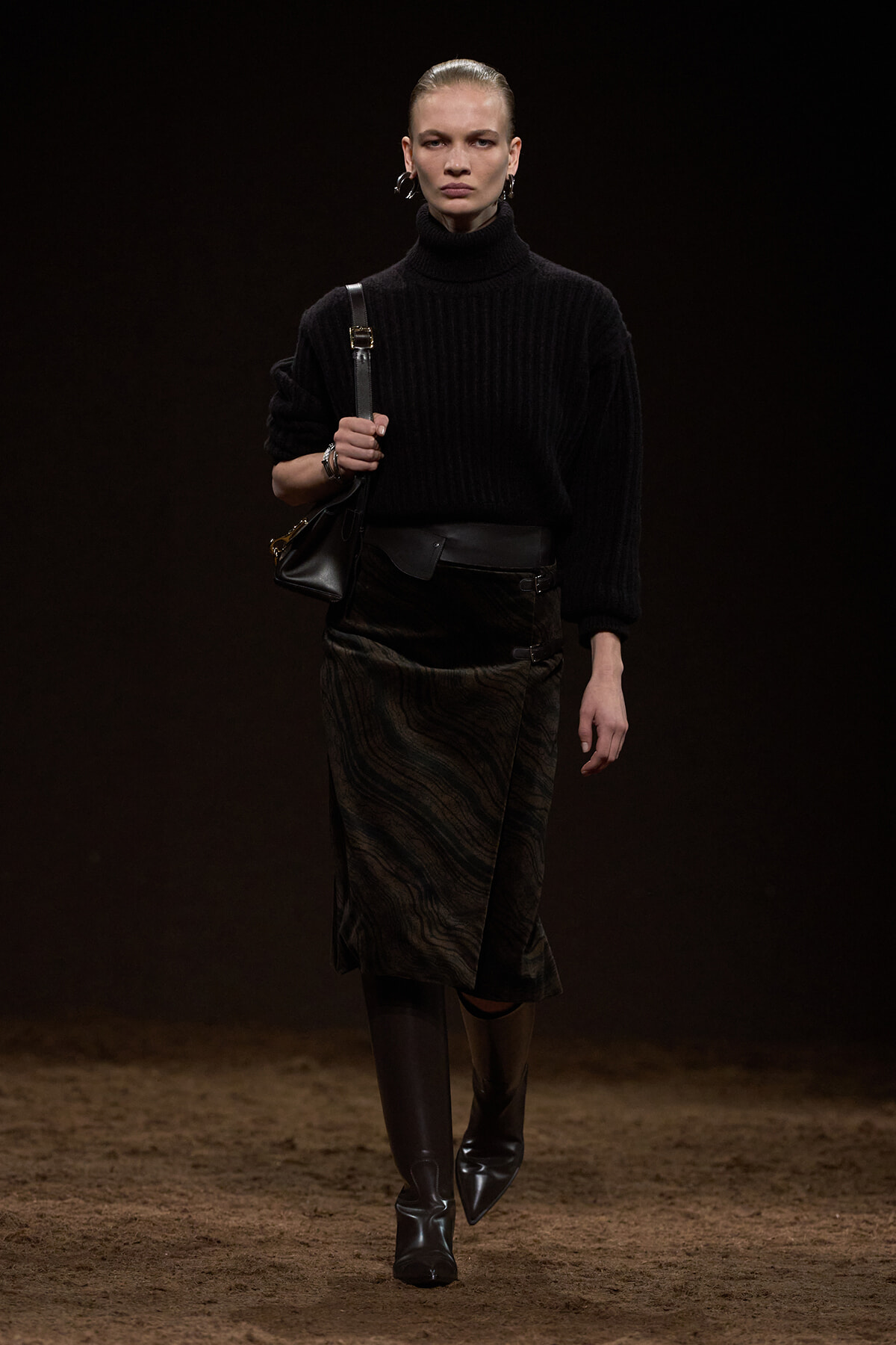 Model walking on a dim runway in a black turtleneck, textured midi skirt, knee-high boots, and a black shoulder bag.