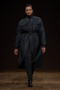 Model walking a runway in a dark quilted trench coat cinched with a wide belt, paired with a black turtleneck and gray leggings.