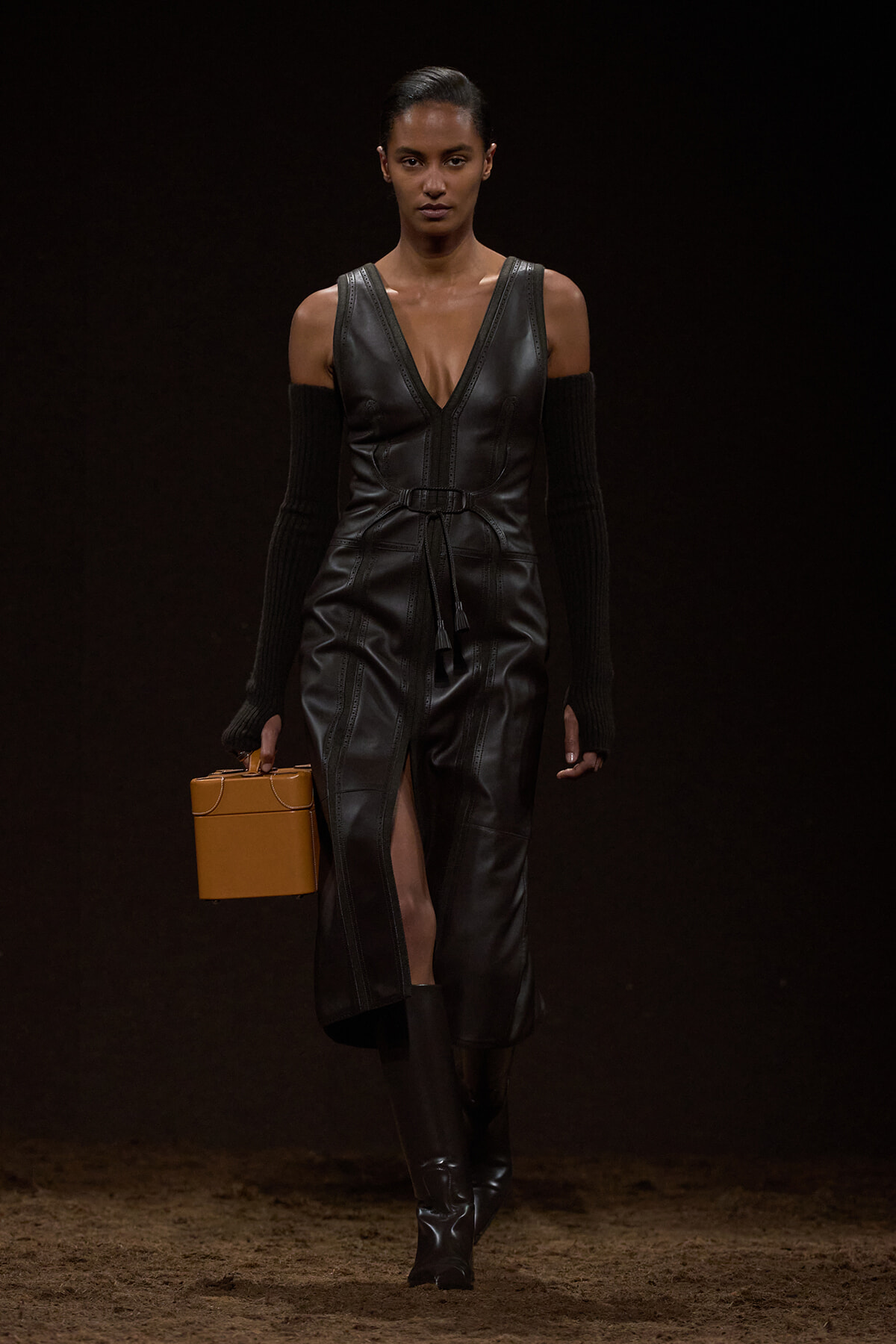 Model on a dark runway wearing a black leather deep-V wrap dress with a belt and thigh-high slit, carrying a tan box clutch