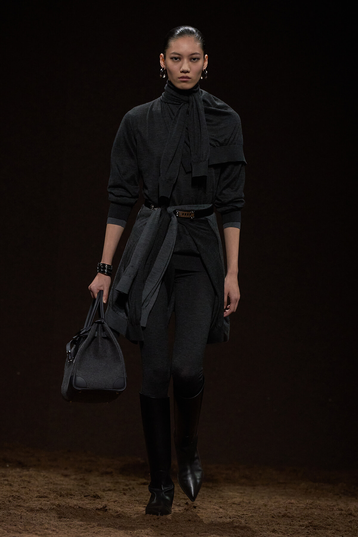 Model walking a runway in a dark charcoal layered outfit with a belt, scarf, and knee-high boots, carrying a gray handbag