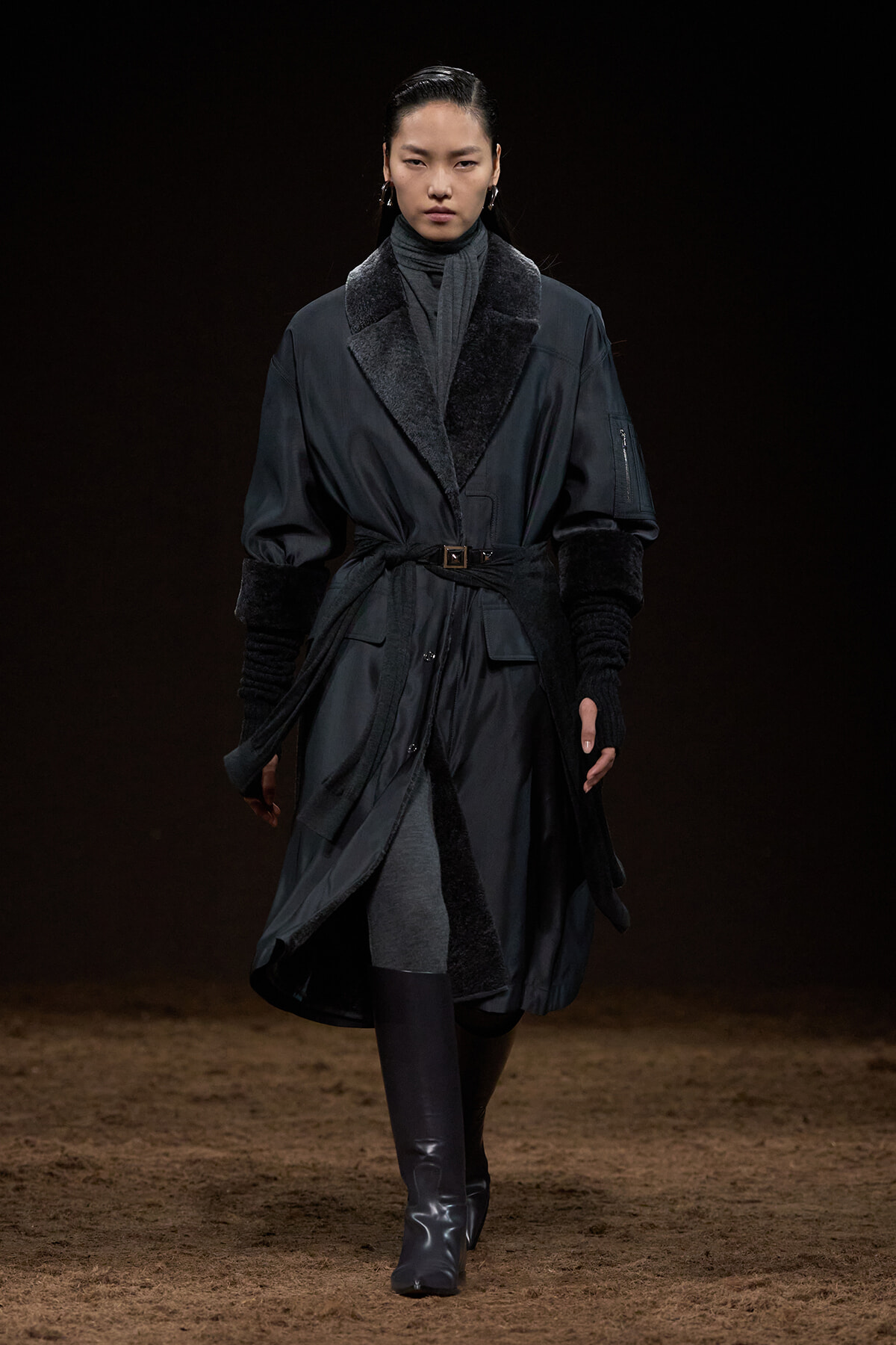 Female model walking a runway in a dark belted coat with fur-trimmed cuffs and collar, pairing with tall boots.