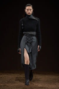 Model on a dim runway wearing a black ribbed turtleneck and a gray wrap skirt with lace-up front, paired with knee-high black boots