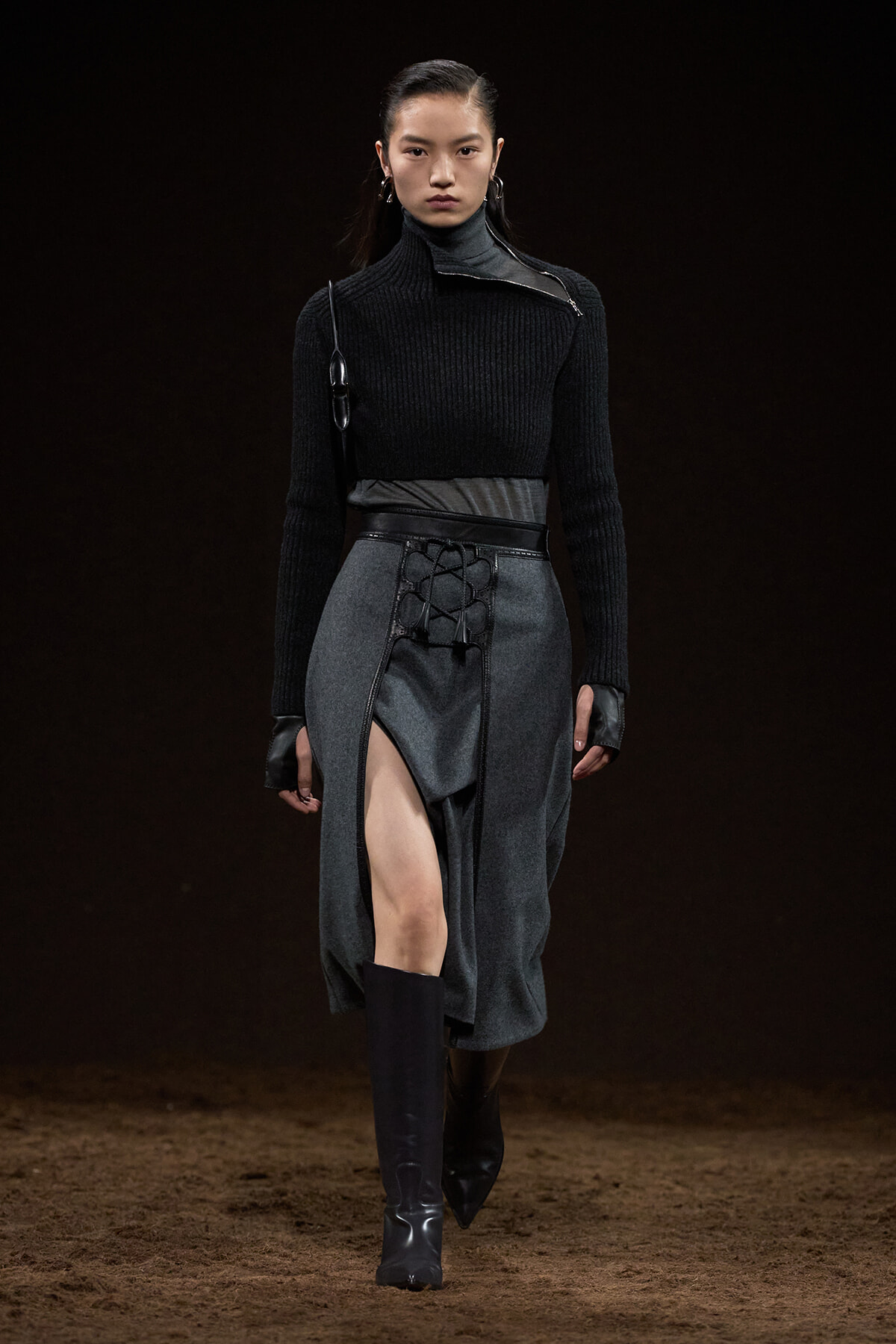 Model on a dim runway wearing a black ribbed turtleneck and a gray wrap skirt with lace-up front, paired with knee-high black boots