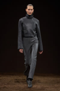 Female model walks a fashion runway in a dark gray leather cropped jacket with an asymmetric zipper and matching pants.