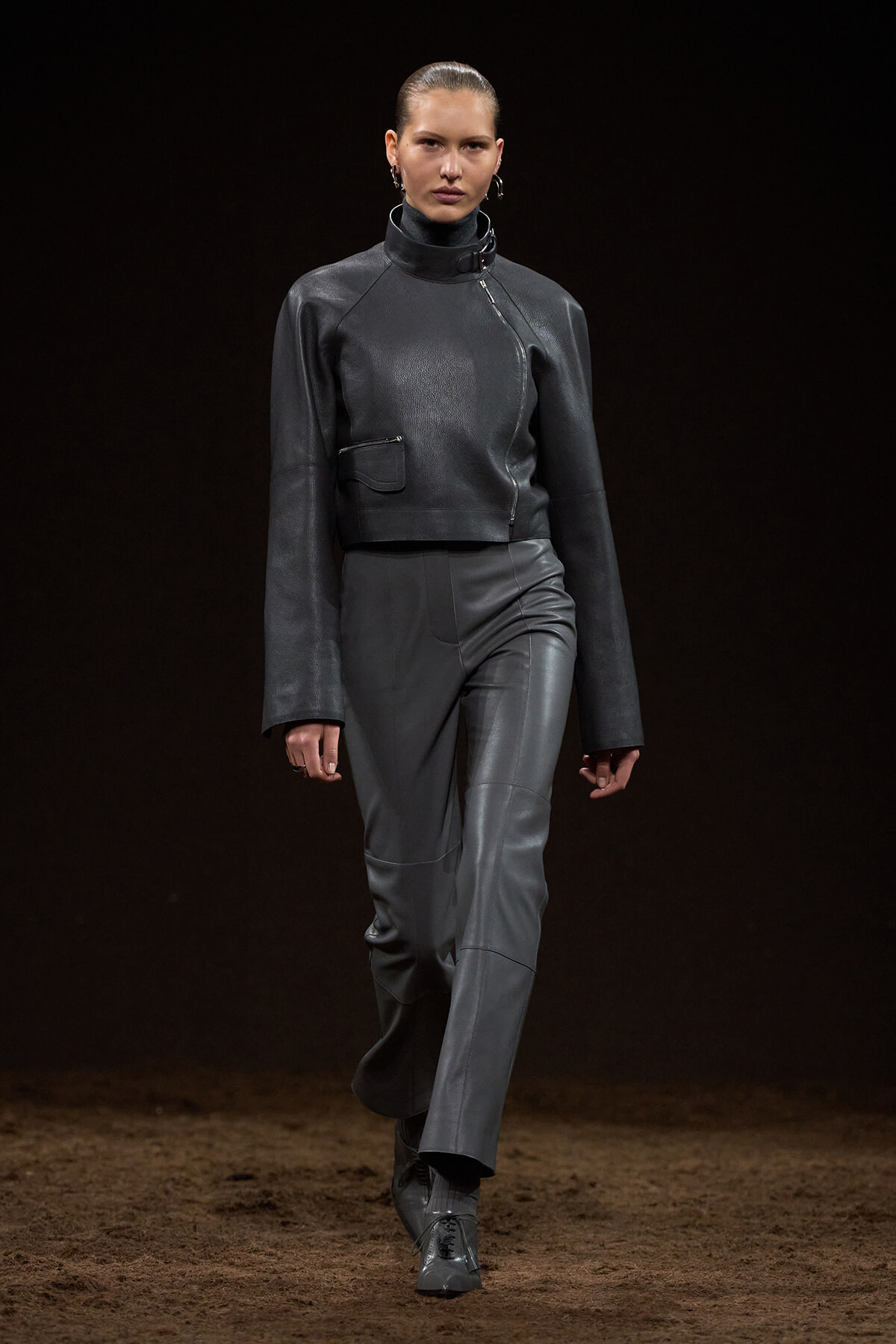 Female model walks a fashion runway in a dark gray leather cropped jacket with an asymmetric zipper and matching pants.