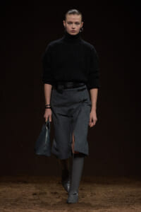 Model on a dark runway wearing a black ribbed turtleneck sweater and a gray wrap-front midi skirt with a front slit, paired with knee-high gray boots and a blue-gray handbag.