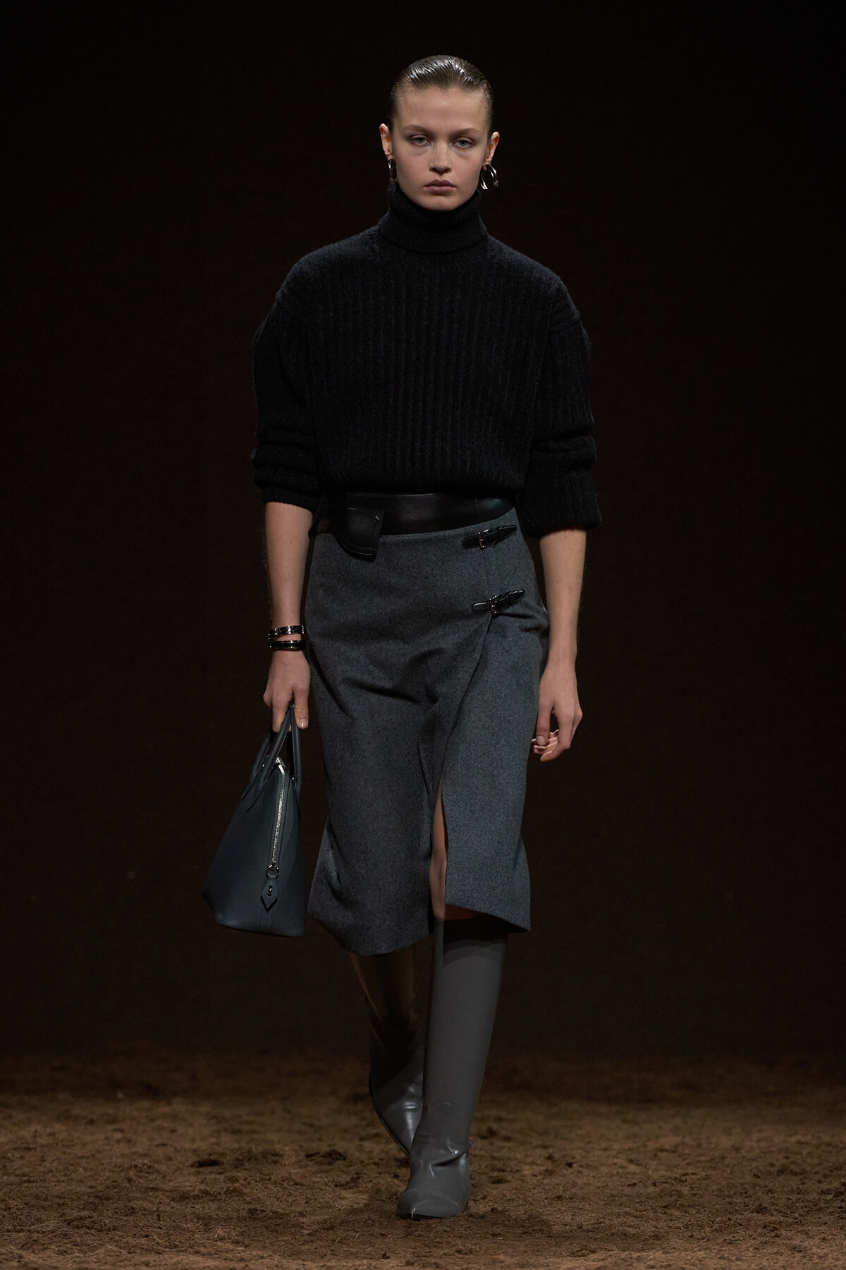 Model on a dark runway wearing a black ribbed turtleneck sweater and a gray wrap-front midi skirt with a front slit, paired with knee-high gray boots and a blue-gray handbag.