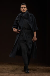 Model walking a fashion run­way in a dark gray layered coat, scarf, and leggings, holding a black clutch.