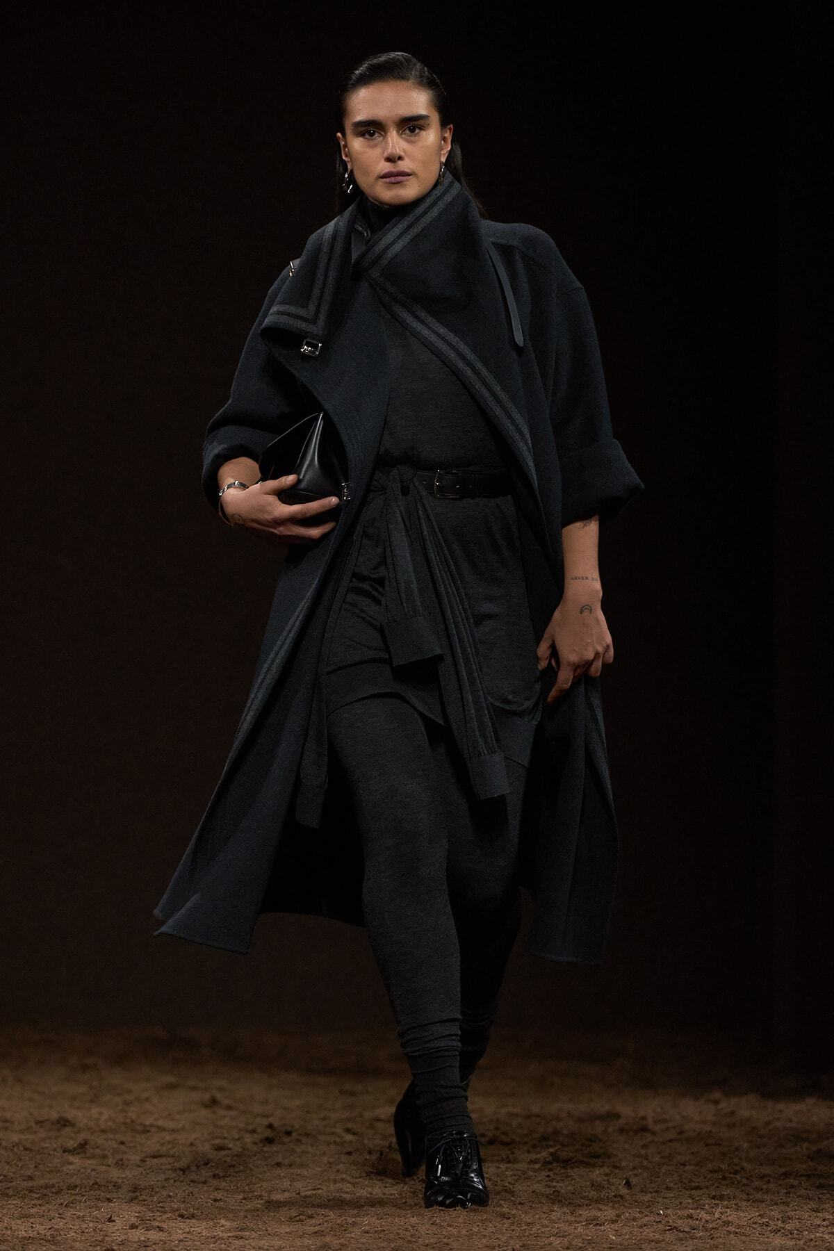 Model walking a fashion run­way in a dark gray layered coat, scarf, and leggings, holding a black clutch.