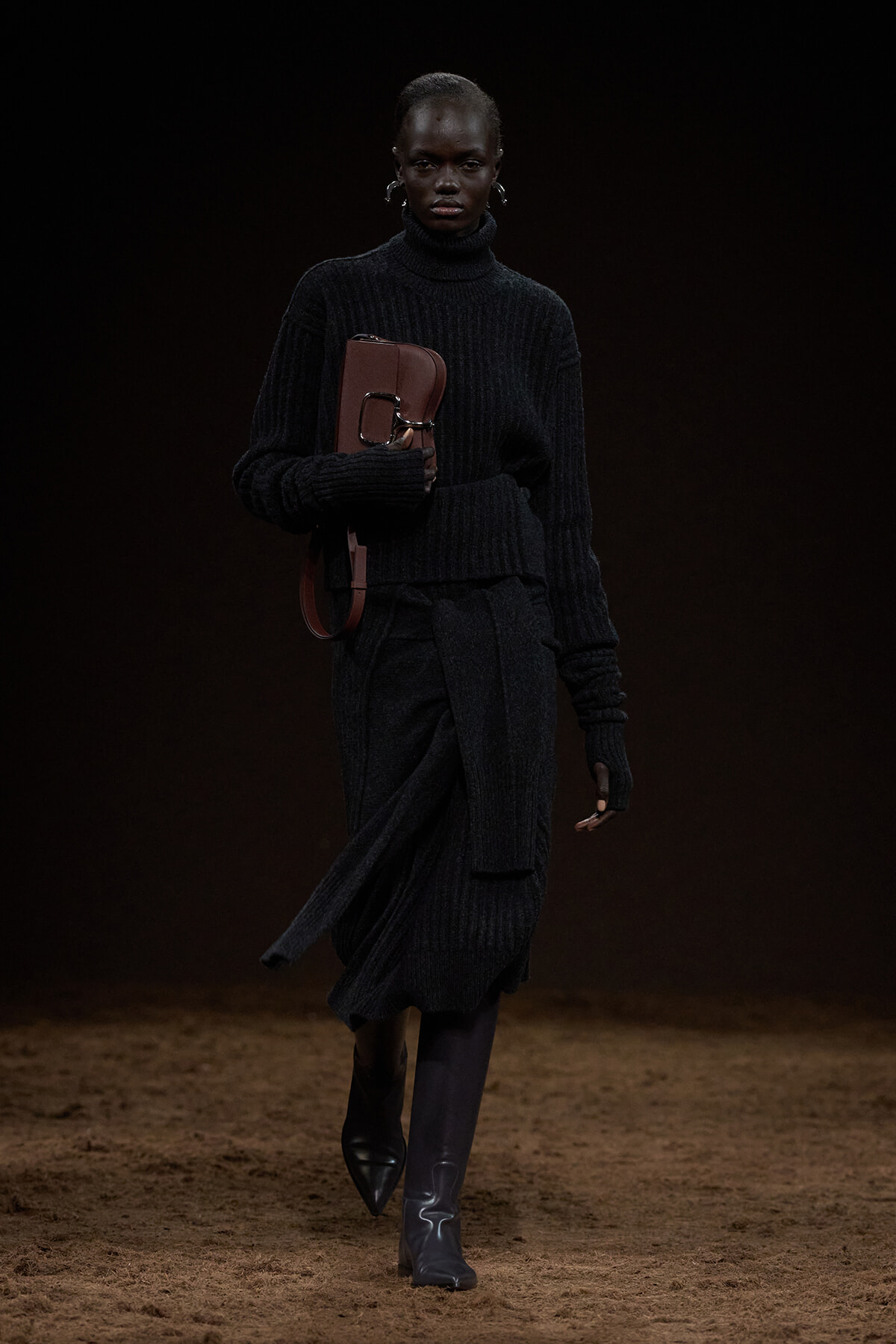 Model walking a fashion runway in an all-black ribbed knit outfit, clutching a brown leather bag with a large buckle.