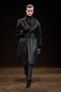 Model walking a dark runway in a black quilted belted coat with a high collar and gloves, carrying a strap over the shoulder.