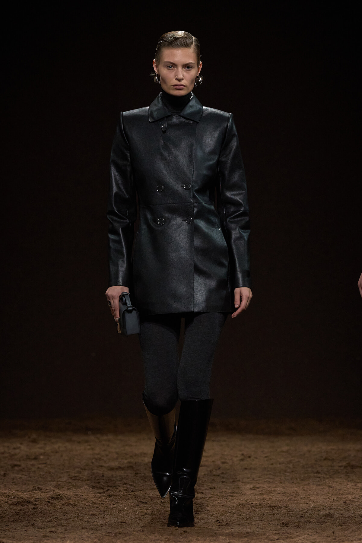 Model on a dark runway wearing a black leather double-breasted coat, gray leggings, and knee-high black boots, holding a small purse