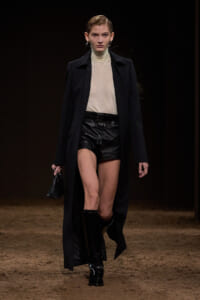 Model walking a fashion runway in a beige turtleneck, black leather shorts, and a long black coat, with knee-high boots and a small black purse.