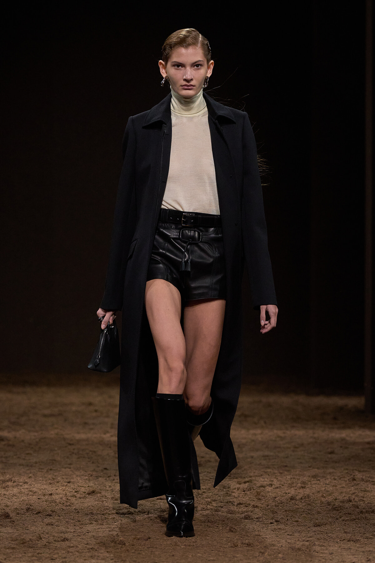 Model walking a fashion runway in a beige turtleneck, black leather shorts, and a long black coat, with knee-high boots and a small black purse.