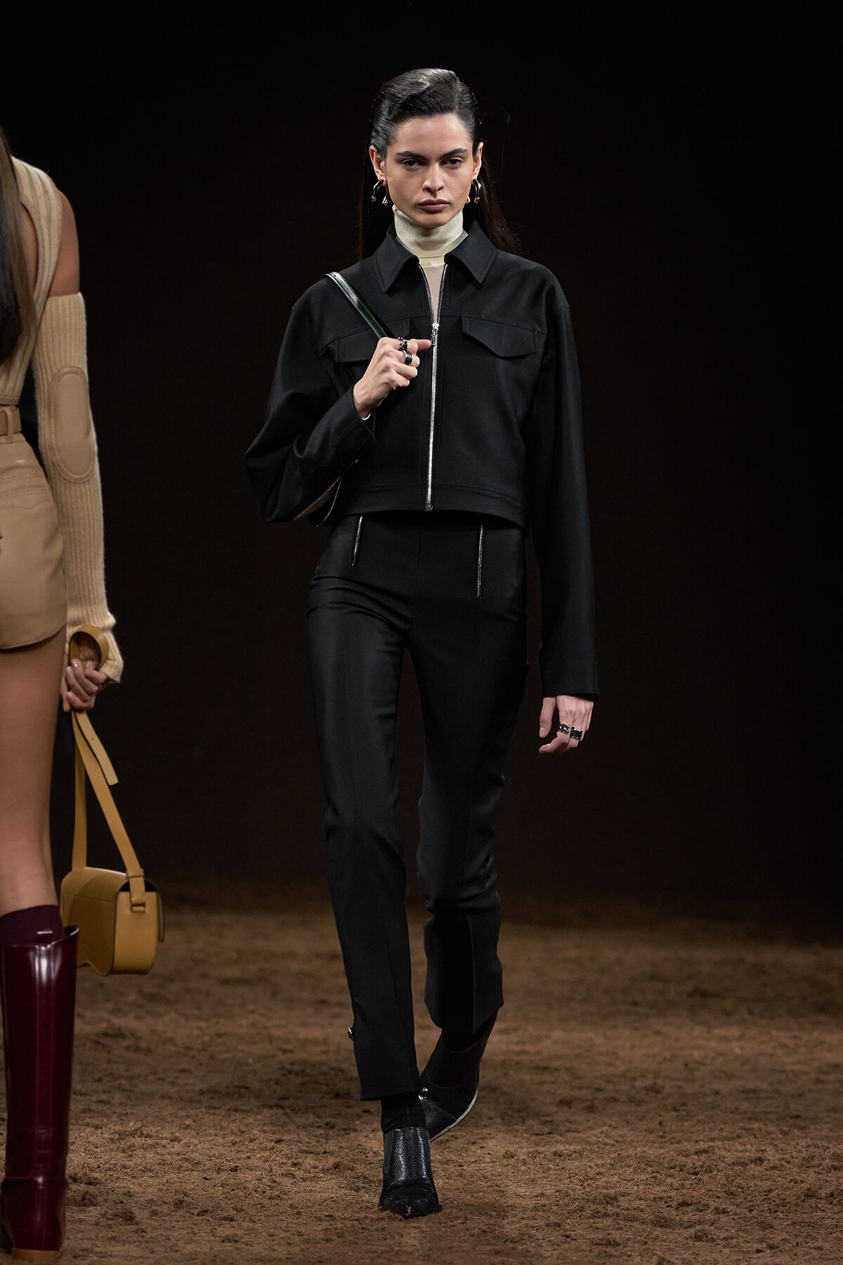 Model walking a fashion show wearing a black cropped jacket with a white turtleneck and black pants, on a brown runway.