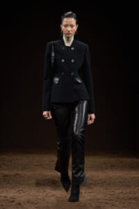 Model on a runway wearing a black double-breasted jacket with leather pants, sleek hair, and a dark backdrop.