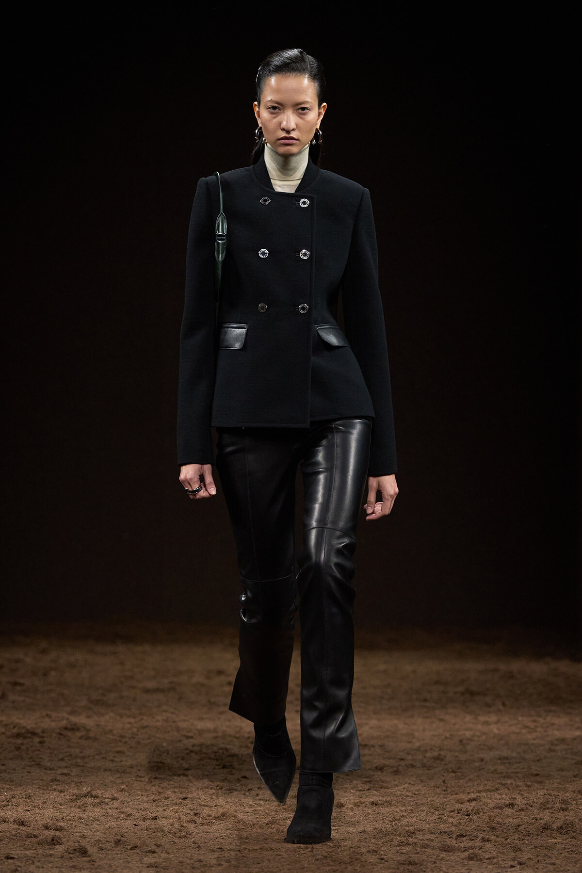 Model on a runway wearing a black double-breasted jacket with leather pants, sleek hair, and a dark backdrop.