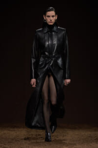 Model walking a runway in a long black leather coat with a cinched waist and wide lapels against a dark background.