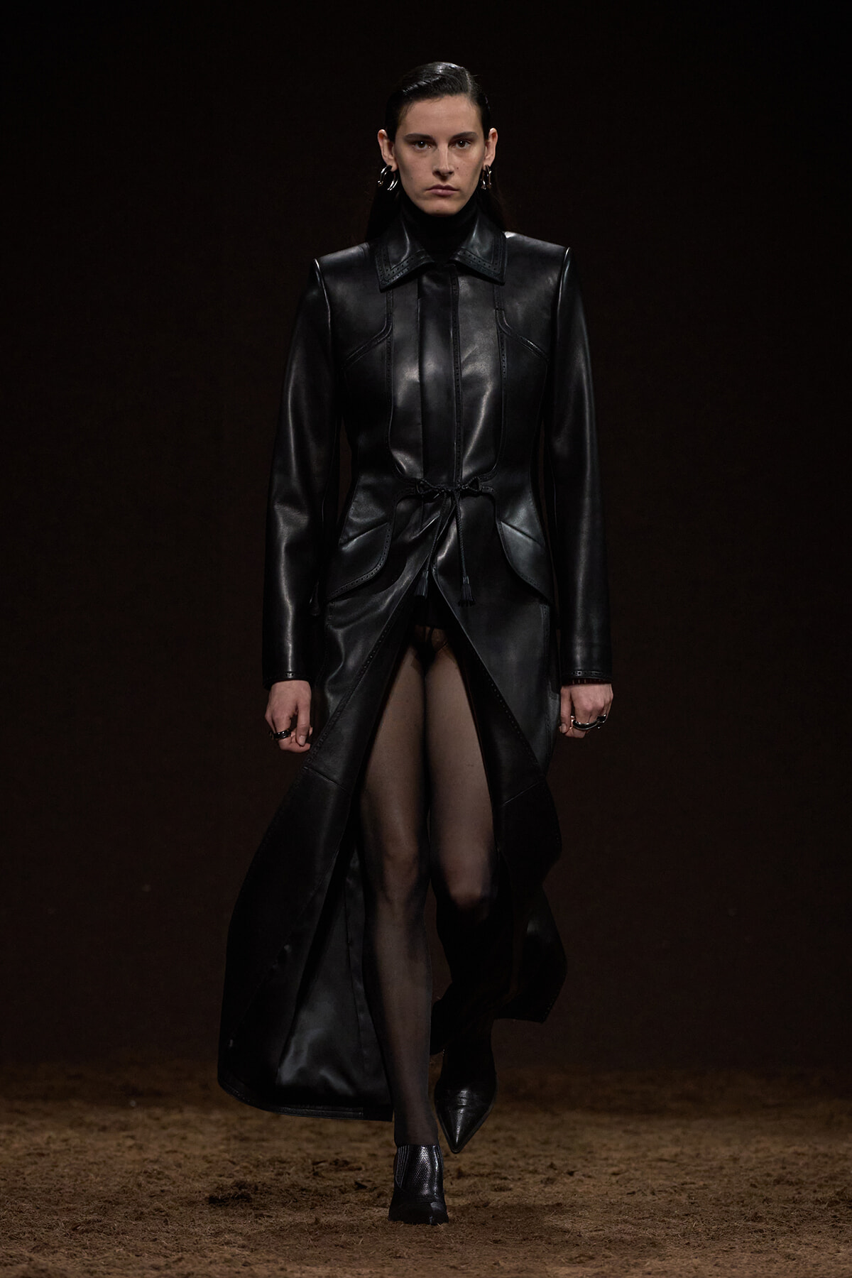 Model walking a runway in a long black leather coat with a cinched waist and wide lapels against a dark background.
