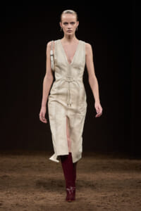 Model walking a runway in a cream sleeveless V-neck belted dress with front slit, paired with burgundy knee-high boots.