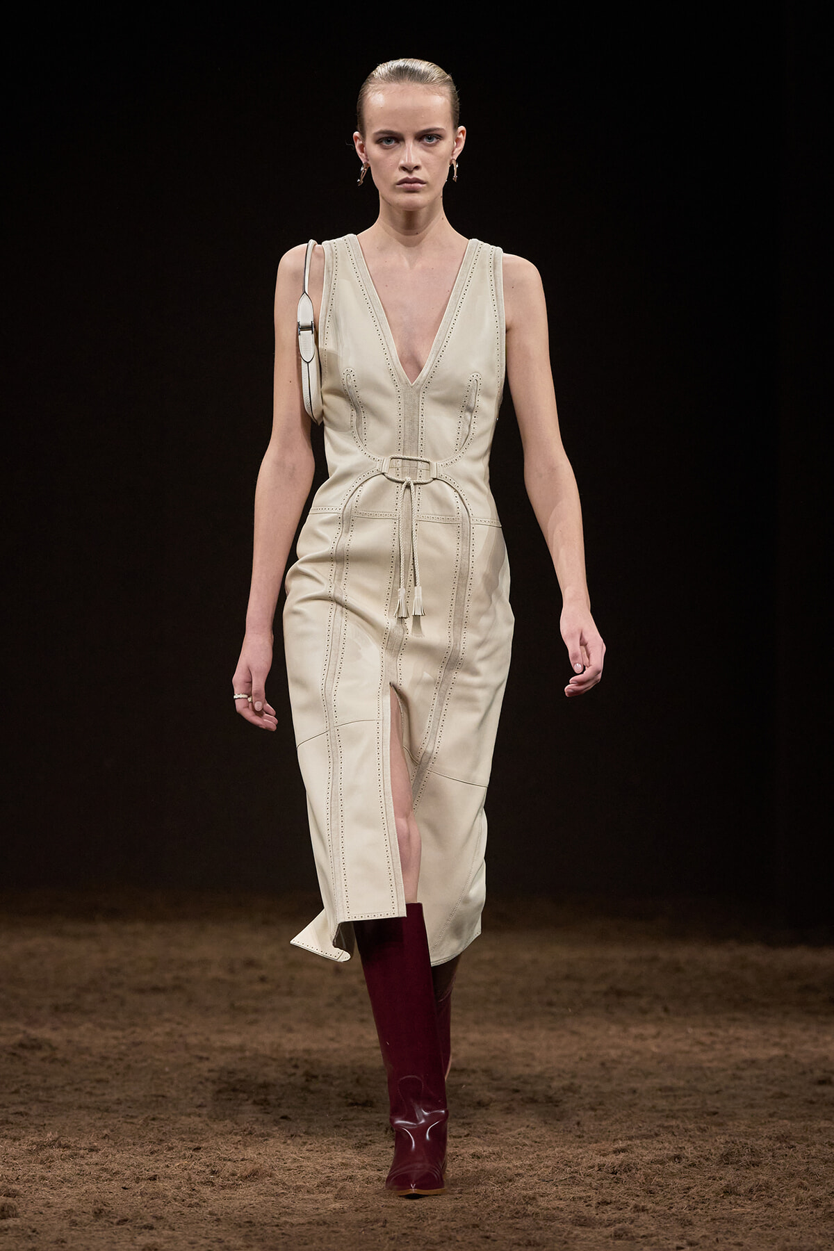 Model walking a runway in a cream sleeveless V-neck belted dress with front slit, paired with burgundy knee-high boots.
