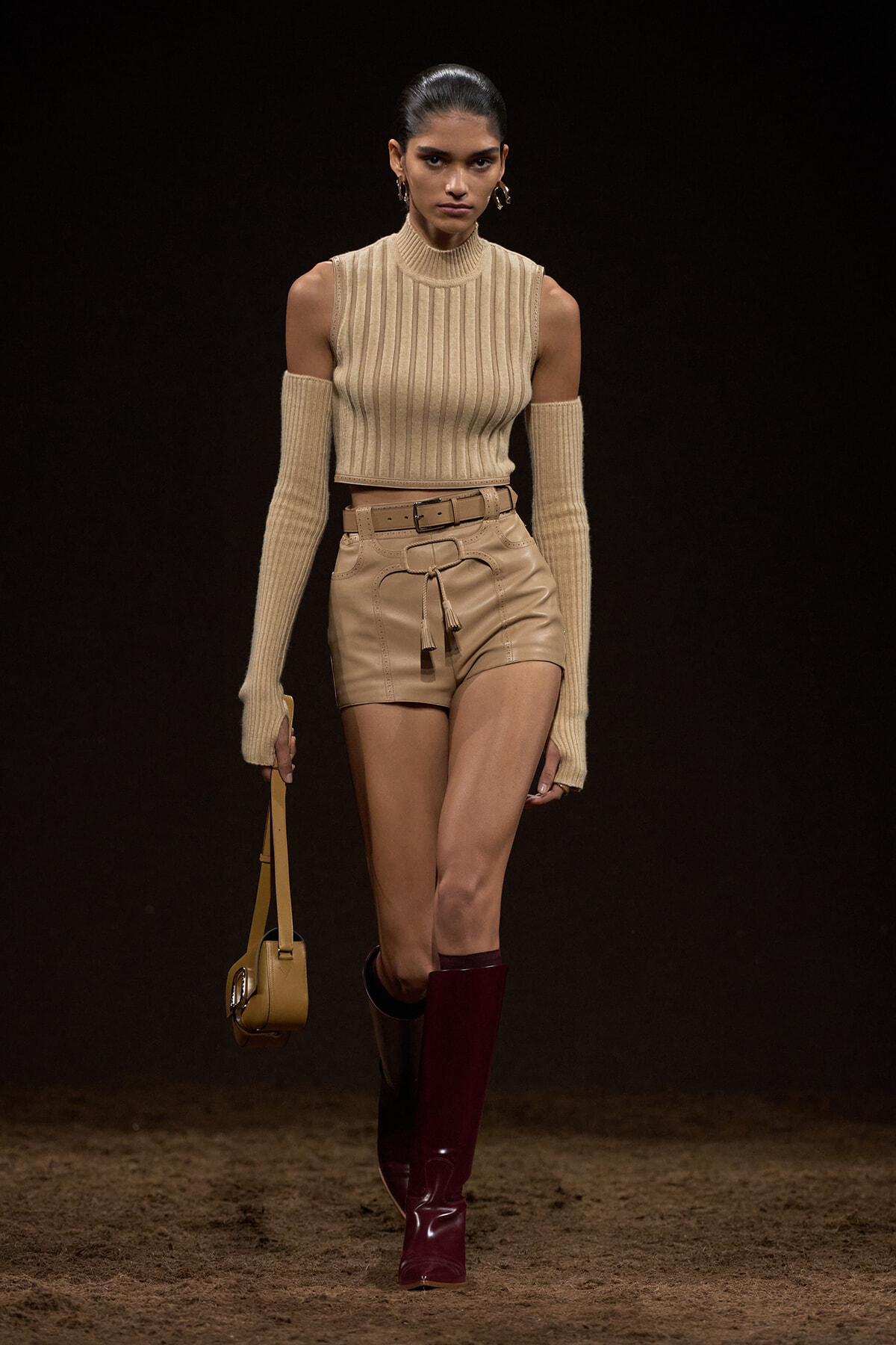 Runway model wearing a beige ribbed sleeveless top with cold-shoulder arms, matching belted beige shorts, burgundy knee-high boots, and a small tan handbag.