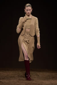 Model walking a fashion runway in a tan coordinated outfit: cropped sweater, sleeveless vest, high-slit lace-up skirt and burgundy boots.