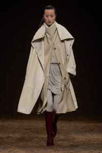 Model on a dark runway wearing an oversized beige trench coat layered over a neutral cream sweater and light gray leggings, with burgundy boots.