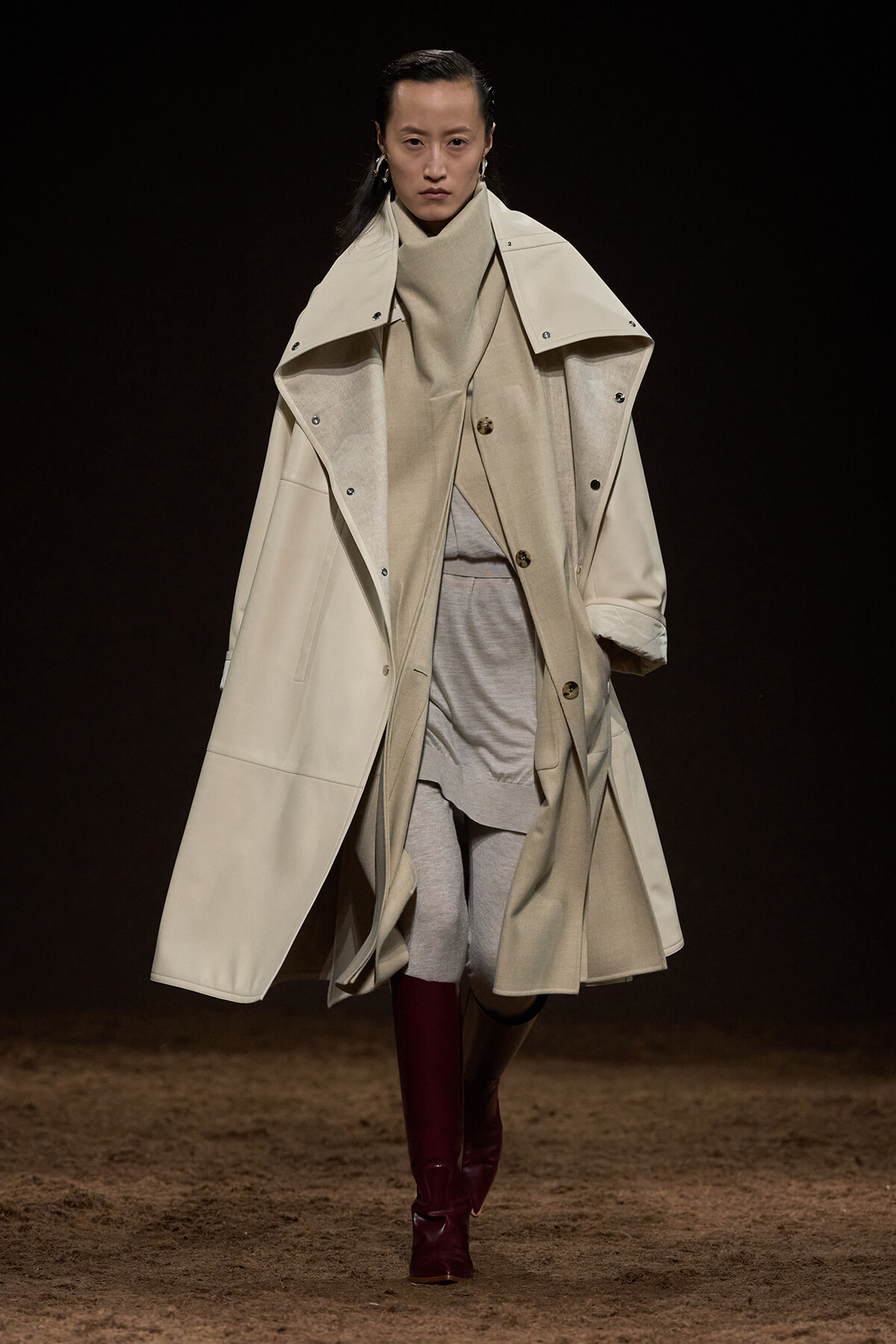 Model on a dark runway wearing an oversized beige trench coat layered over a neutral cream sweater and light gray leggings, with burgundy boots.