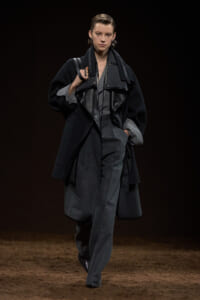 Model walking a runway in a dark, monochrome ensemble: oversized black coat with grey lining over matching trousers and a leather-panel jacket.",