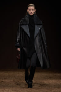 Model on a dim runway wearing a long black leather coat over a black turtleneck, carrying a small brown handbag at her side.