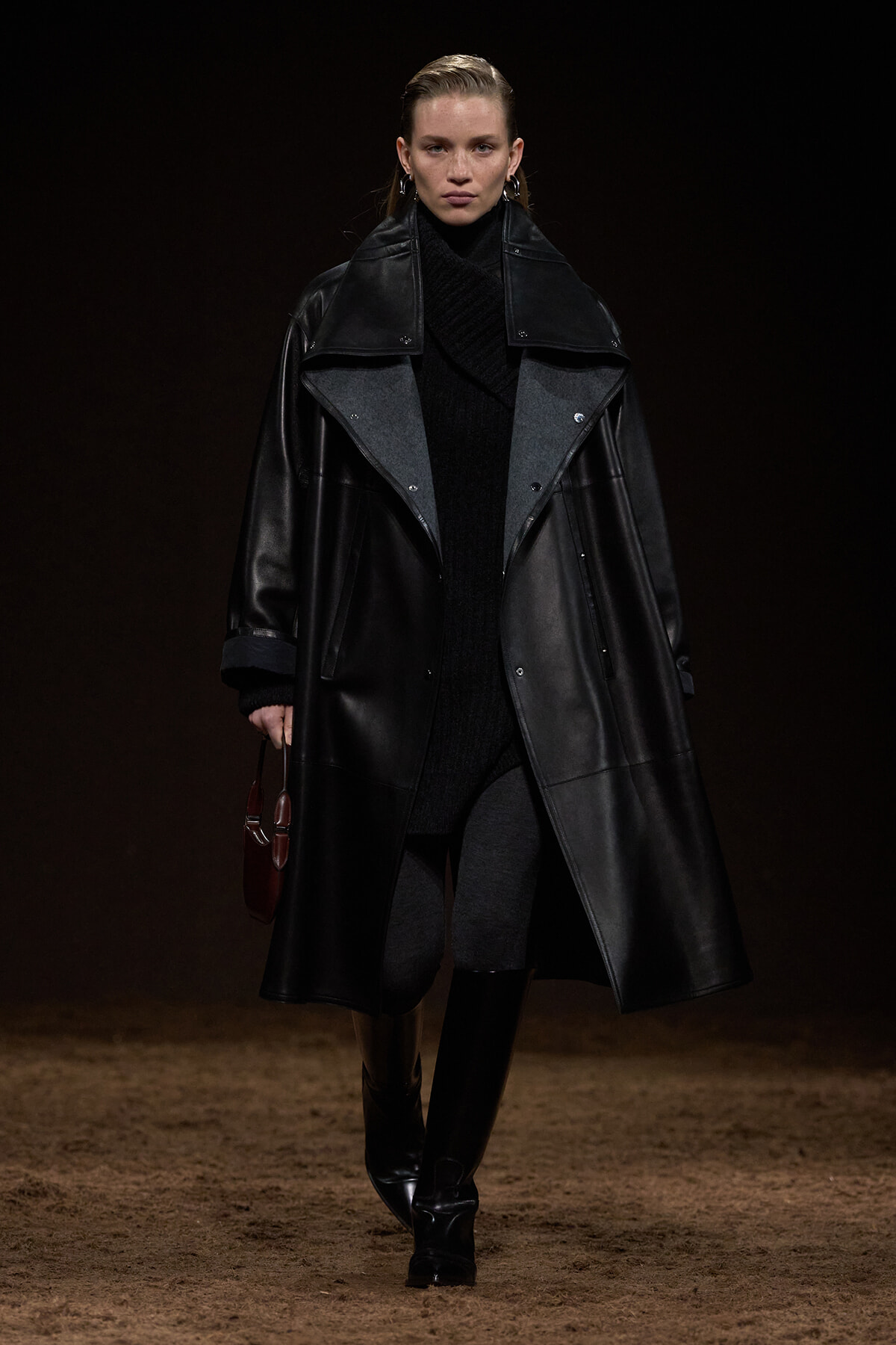 Model on a dim runway wearing a long black leather coat over a black turtleneck, carrying a small brown handbag at her side.