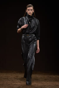 Model walking a fashion runway in an all-black leather outfit: a high-neck top with wide-leg pants, belt, and shoulder bag, against a dark backdrop.