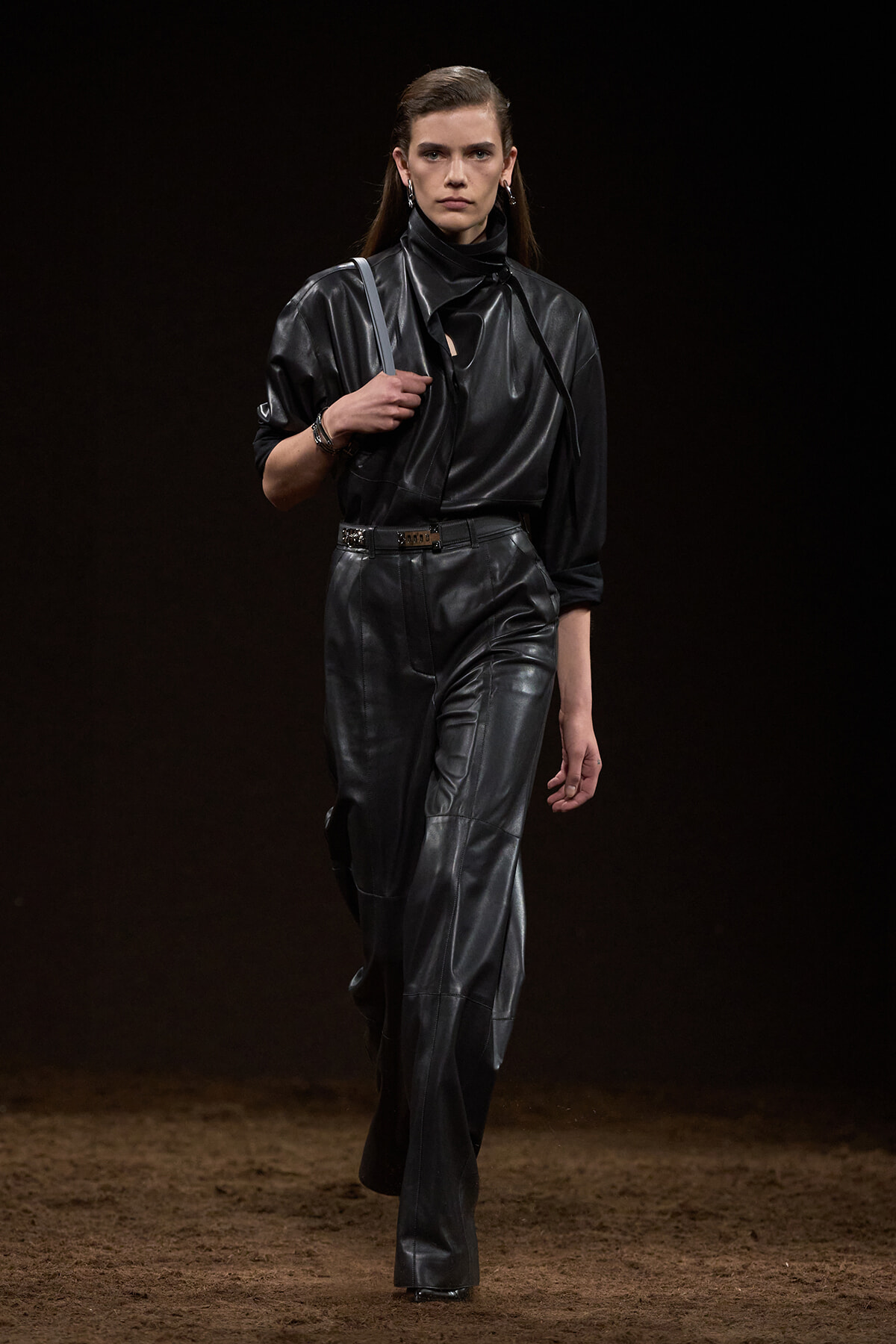 Model walking a fashion runway in an all-black leather outfit: a high-neck top with wide-leg pants, belt, and shoulder bag, against a dark backdrop.