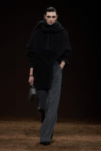 Model walking a fashion runway in an all-dark ensemble: oversized black knit with a large scarf, wide‑leg gray trousers, and a small structured gray handbag.