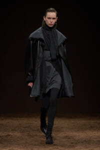 Model walking the runway in a monochrome, layered black and gray outfit featuring an oversized leather coat, cape, scarf, belt, and tall lace-up boots.
