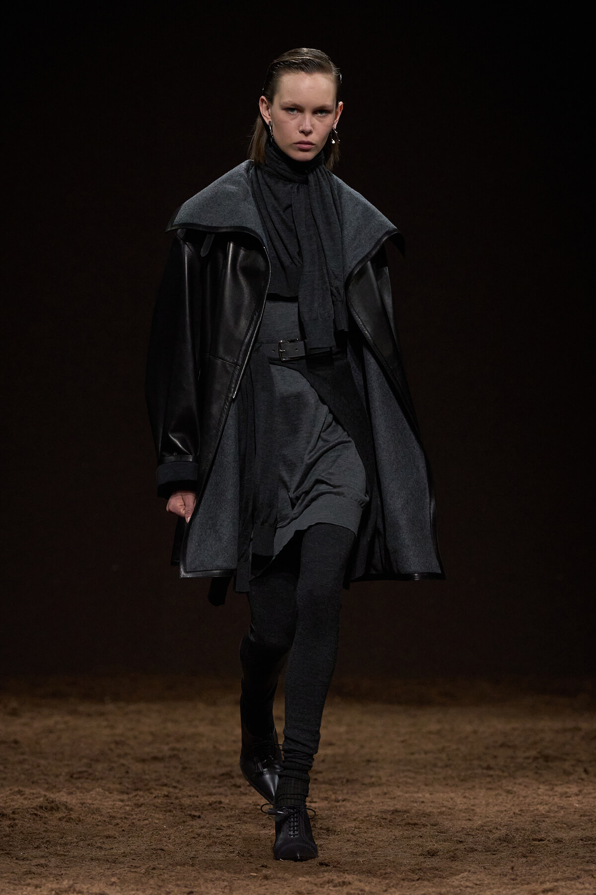 Model walking the runway in a monochrome, layered black and gray outfit featuring an oversized leather coat, cape, scarf, belt, and tall lace-up boots.