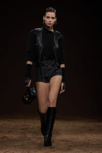 Model walking a fashion runway in an all-black outfit: leather vest, knit sweater, black shorts, knee-high boots, and a black handbag.