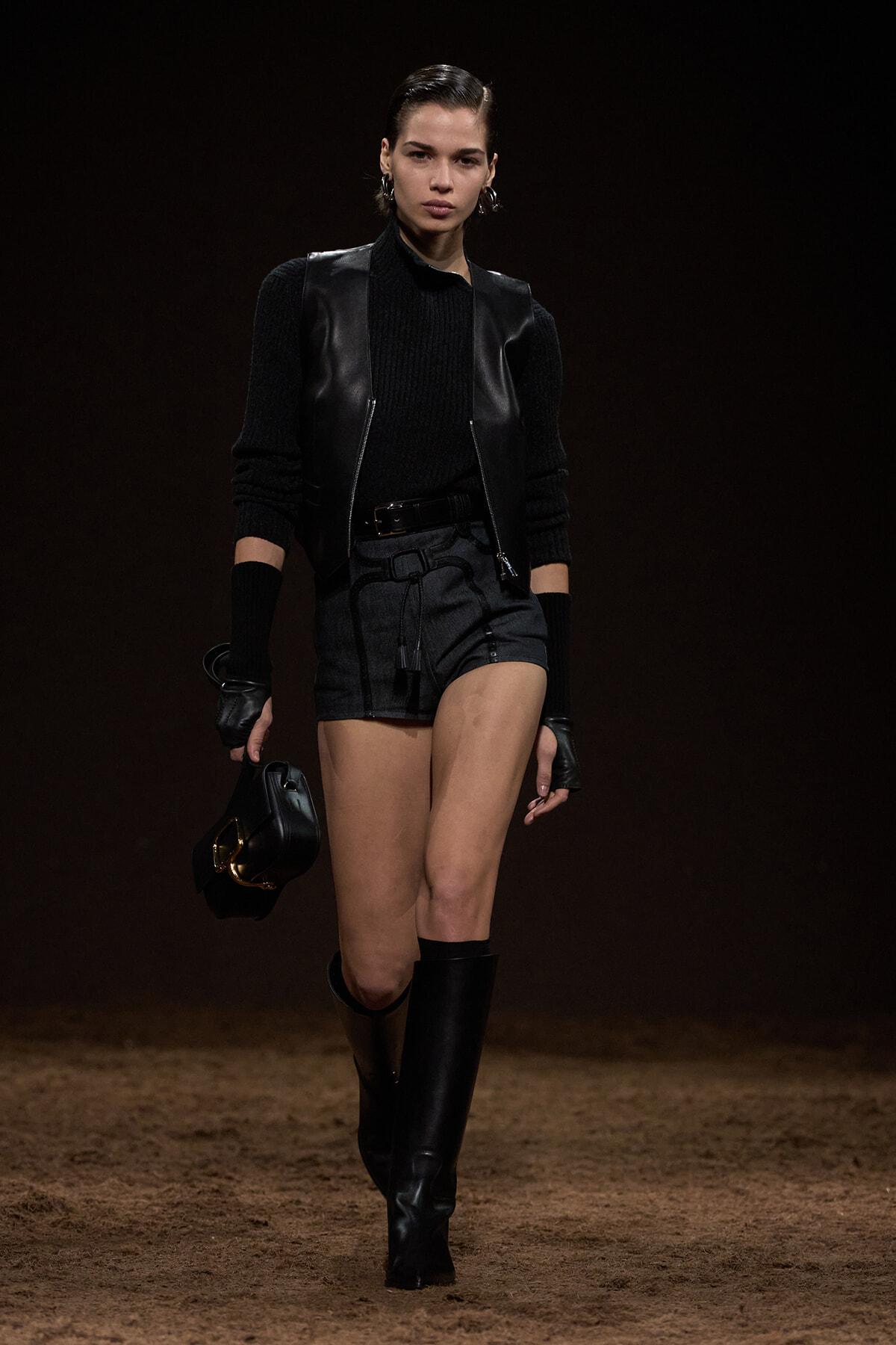 Model walking a fashion runway in an all-black outfit: leather vest, knit sweater, black shorts, knee-high boots, and a black handbag.